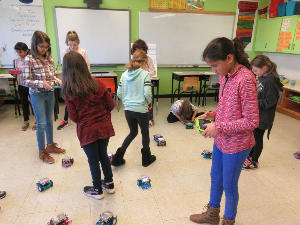 Young students are in a classroom with robots on the ground. They are using tablets to control them.