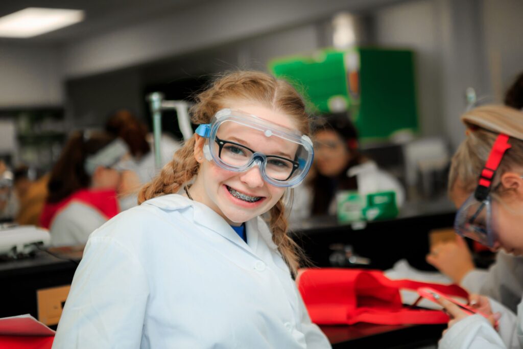 A young girl wearing safety glasses and a white protective suit. She is smiling at the camera.